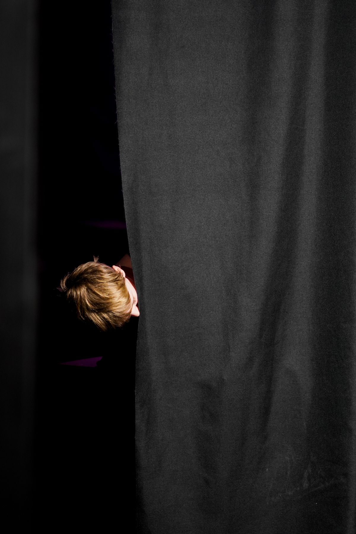 A young performer peeking out from behind a black stage curtain